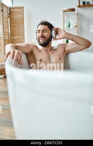 joyful attractive man with beard and headphones sitting and relaxing in his bathtub, mental health Stock Photo