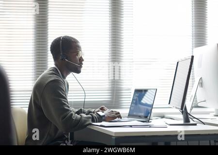 Side view portrait of Black young man wearing headset and using computers as client support operator IT company copy space Stock Photo