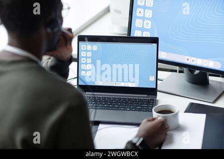 Over shoulder view of young Black man using laptop with blue desktop screen in minimal office setting and drinking coffee copy space Stock Photo