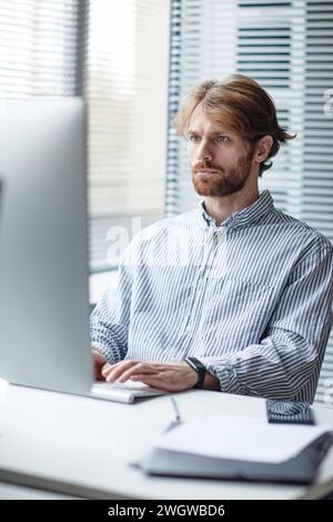 Graphic vertical portrait of bearded adult man using computer at workplace in IT office against window Stock Photo
