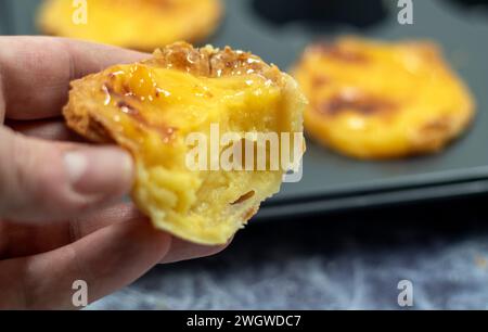 A man's hand holds a half-eaten bitten fresh Portuguese Pastel de Nata pie with custard on the background of a baking dish. Pastel de Belem is a pastr Stock Photo