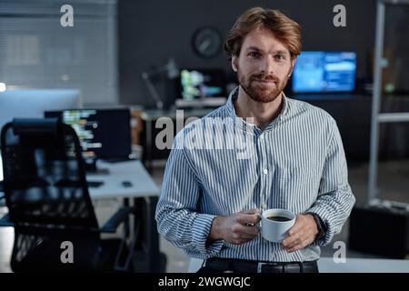 Waist up portrait of smiling bearded man holding coffee cup and looking at camera standing in formal IT office with blue neon light copy space Stock Photo