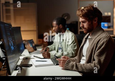 Side view portrait of bearded man using smartphone at desk while working late in dark office, copy space Stock Photo