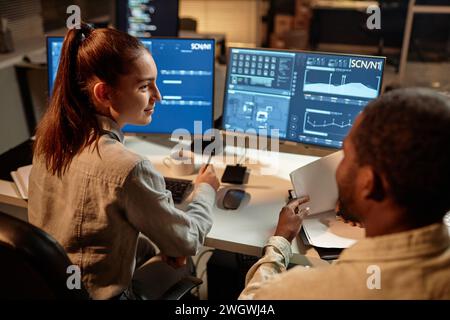 Side view portrait of woman with male colleague looking at computer screen with data in dark office Stock Photo