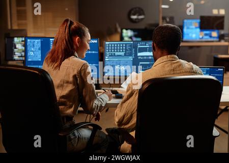 Back view at two IT developers man and woman looking at computer screen with data in dark office Stock Photo