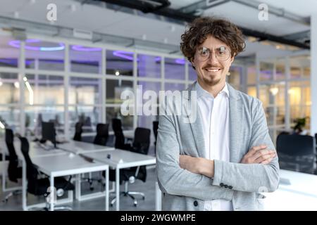 Portrait of a cheerful young businessman with curly hair confidently standing in a contemporary office space, arms crossed. Stock Photo