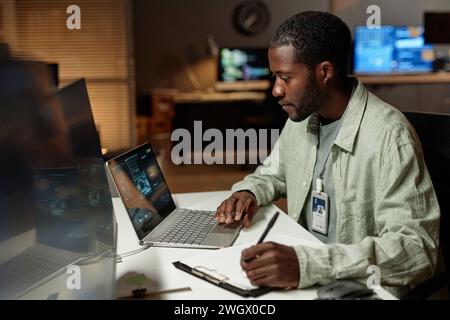 Side view portrait of Black young man using computer and taking notes while working late in dark office, copy space Stock Photo