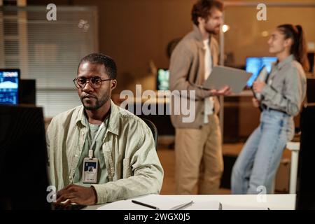 Portrait of Black computer programmer wearing glasses working in office at night with people in background, copy space Stock Photo
