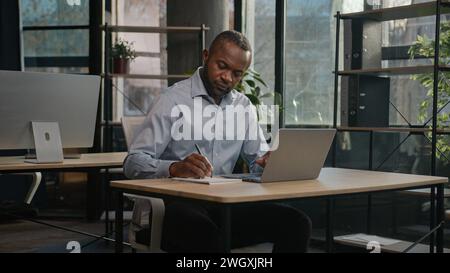 Mature adult 40s middle-aged businessman African American ethnic man journalist manager writing text in paper notebook typing on laptop write notes Stock Photo