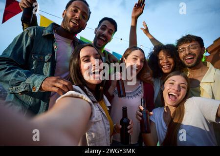 Joyful attractive young woman taking selfie at millennial alcohol party surrounded by group friends. Stock Photo