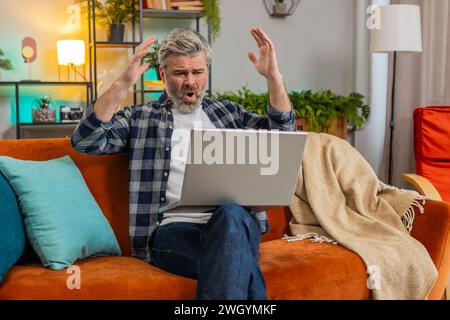 Upset senior man with gray hair sitting on sofa at home uses laptop having anxiety and stress. Shocked Caucasian mature middle-aged guy reacting to loss bad news on netbook in living room in apartment Stock Photo
