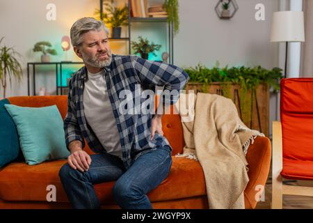 Tired senior bearded man suffering from back pain sitting on couch in living room at home. Sad old male massages painful lower back muscles. Caucasian mature guy exhausted overworked in apartment. Stock Photo