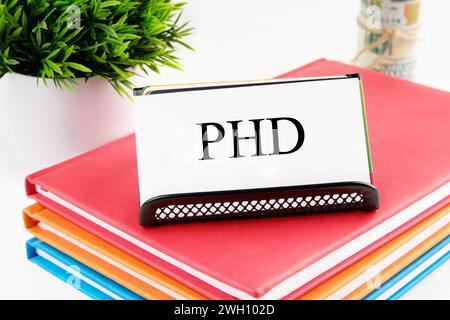 PHD text on a white business card standing on a stand on notebooks, next to the money out of focus and a green plant on a white background Stock Photo