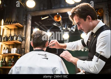 Barber giving haircut to young man in barbershop. Professional barber well-dressed in vest and white shirt with rolled-up sleeves, concentrating on cutting the hair, using comb. Stock Photo