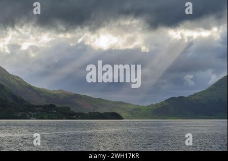 Sunbeams break through the dramatic cloud cover, illuminating parts of a serene lake surrounded by mountains as evening approaches. Stock Photo
