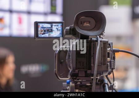 A high-definition television camera equipped with a monitoring screen is capturing a live stream at a busy exhibition center, focusing on an unidentif Stock Photo