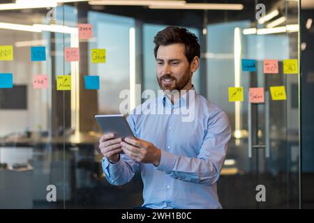 Mature businessman reviewing digital data on a tablet with sticky notes on glass wall in a well-lit contemporary office. Stock Photo