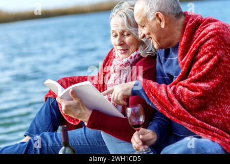Happy senior couple enjoying time together reading a book by the lake wrap around in a red blanket. Stock Photo