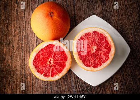 Red grapefruit sliced on a white plate, sitting on a dark wooden table. Healthy eating. Fruits in the kitchen. Citrus and vitamins. Vegan diet. Stock Photo