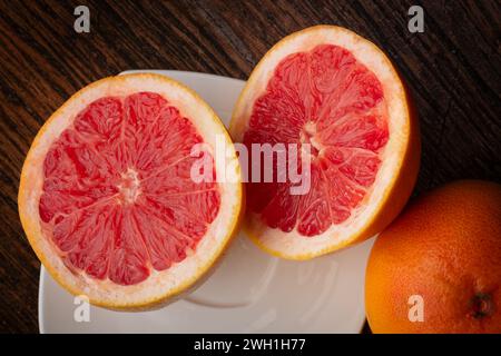Red grapefruit sliced on a white plate, sitting on a dark wooden table. Healthy eating. Fruits in the kitchen. Citrus and vitamins. Vegan diet. Stock Photo