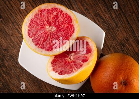 Red grapefruit sliced on a white plate, sitting on a dark wooden table. Healthy eating. Fruits in the kitchen. Citrus and vitamins. Vegan diet. Stock Photo
