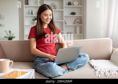 Relaxed young asian woman smiling while browsing on her laptop Stock Photo