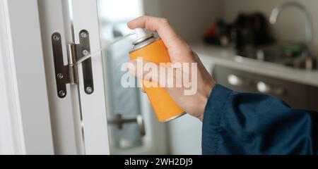 closeup of a caucasian man, in blue overalls, spraying some lubricating oil on the hinge of a white door, in a panoramic format to use as web banner o Stock Photo