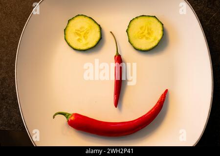 Smilng face arranged on a plate with red chili peppers and slices of snake cucumber. Stock Photo