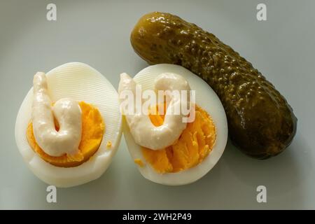 Close up of two eggs cut in halves dressed with mayonnaise and a cucumber on a white grey background. Stock Photo
