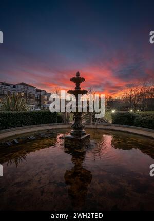 Fountain and pond in a park at sunset with a pink sky. Evening view in portrait orientation with buildings and trees behind. Stock Photo