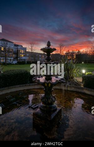 Fountain and pond in a park at sunset with a pink sky. Dusk view in portrait orientation with buildings and trees behind. Stock Photo