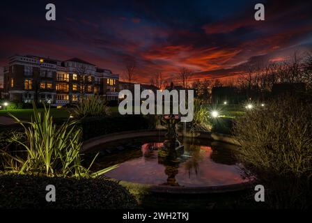 Fountain and pond in a park at sunset with a red sky. Night view in landscape orientation with buildings and trees behind. Stock Photo