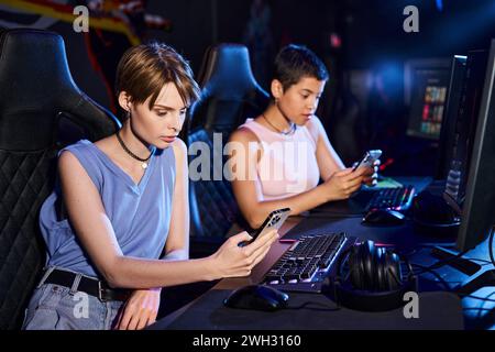 Two young women sit at computer desk while scrolling their phones in cybersport game club Stock Photo