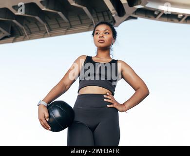 Young woman in black fitness attire under a bridge taking a break. Female athlete with medicine ball outdoors looking away. Stock Photo