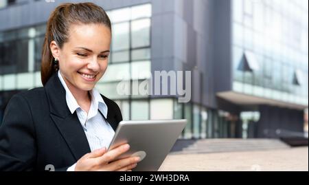 Young brunette businesswoman looking at digital tablet and smiling while standing on the street in front of business building. Stock Photo