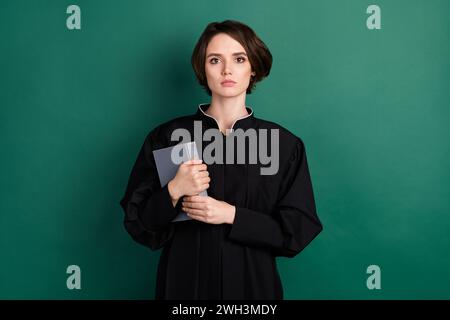 Photo of serious cool confident woman judge attorney wearing black robe holding book isolated on green color background Stock Photo