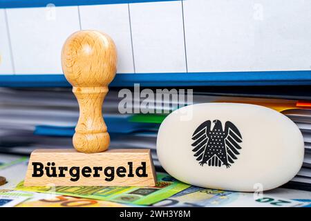 February 7, 2024: A stamp next to folders and euro banknotes with a logo of the German federal eagle with the inscription: Bürgergeld.FOTOMONTAGE *** Ein Stempel neben Ordnern und Euro Geldscheinen mit einem Logo des deutschen Bundesadlers mit der Aufschrift: Bürgergeld. FOTOMONTAGE Stock Photo