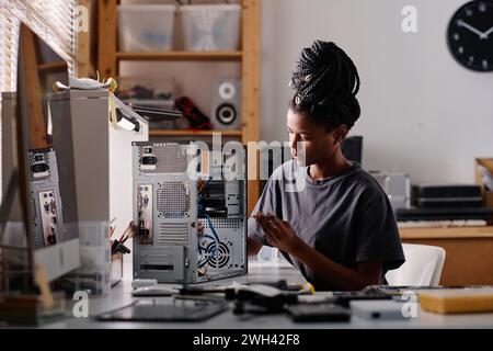 Black female technician sitting at table in workshop and repairing system unit using screwdriver Stock Photo