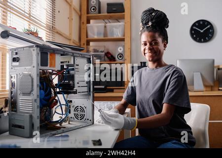 Young black woman sitting at her table and smiling at camera while repairing computer system unit Stock Photo