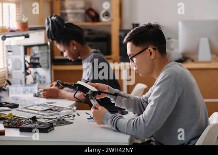 Young technician sitting at table and fixing faulty drone, his black colleague working on background Stock Photo