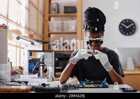 Female african american technician wearing glasses with binocular loupes attentively examining CPU Stock Photo