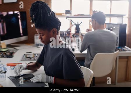 Black woman with braids repairing cellphone at workshop desk, her coworker sitting on background Stock Photo