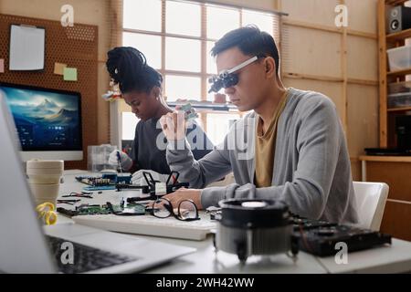 Young repairman in glasses with loupes examining CPU sitting next to his black female colleague Stock Photo