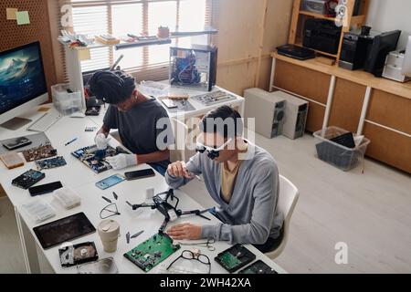 Biracial technicians sitting side by side at table in workshop and fixing faulty electronic devices Stock Photo