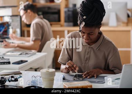 Black technician repairing broken phone with screwdriver, her male colleague working on background Stock Photo