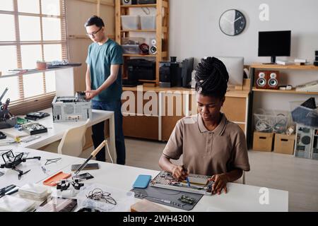 Black technician soldering element of electronic device, her male colleague working on background Stock Photo