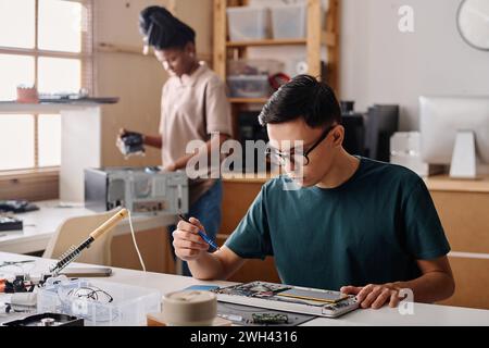Young technician using wireless soldering iron for work, his black colleague standing on background Stock Photo