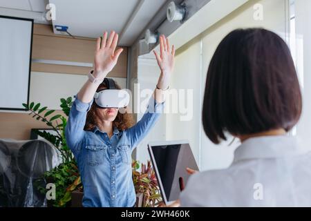 woman with curly brown hair and denim shirt trying on VR in the office with asian colleague in front working with tablet Stock Photo