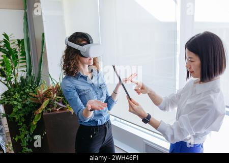 two standing female colleagues in the office, one with curly hair wearing VR and another with short dark hair holding tablet Stock Photo