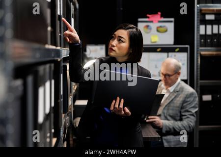 Asian private detective in criminal cases archive office looking for criminology file paperwork on cabinet shelves. Investigator woman surrounded by criminal case folders in agency repository room Stock Photo